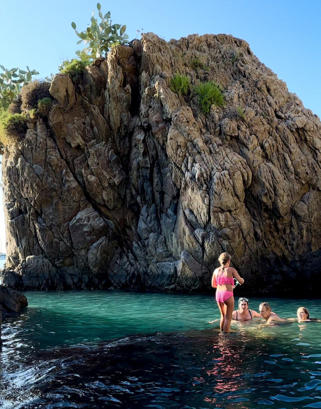 a cove with a huge bolder and people swimming and a little girl standing on the edge of a submerged rock