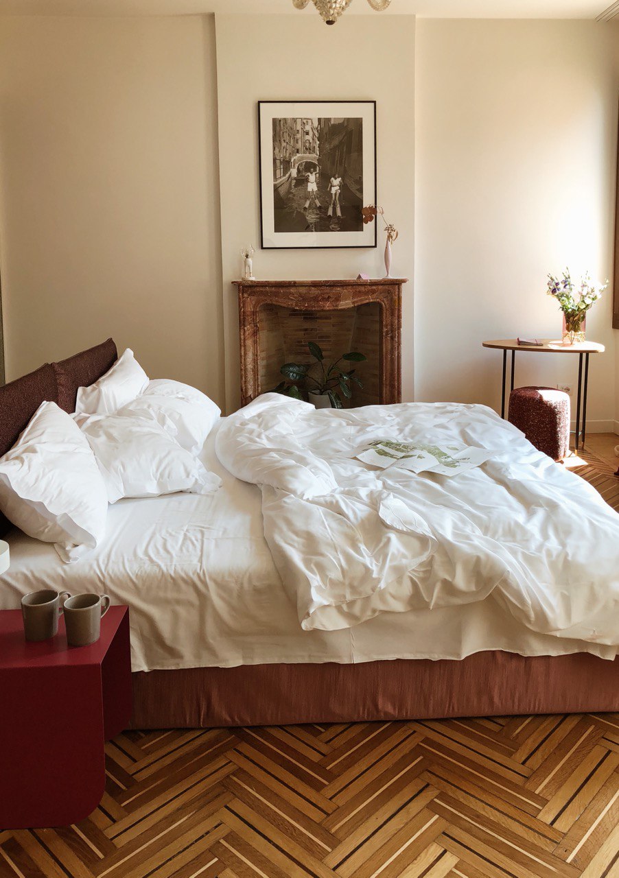 Casa Flora - an unmade large bed with white sheets and a fireplace with a large plant inside and two mugs on the bedside table
