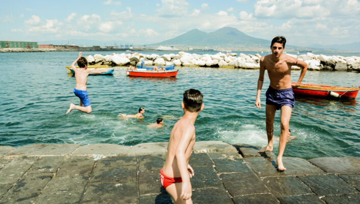 an image by Francesco Freddo of young boys jumping off of the concrete ledge into the clear blue water in the cove off of Naples under Mt Vesuvius