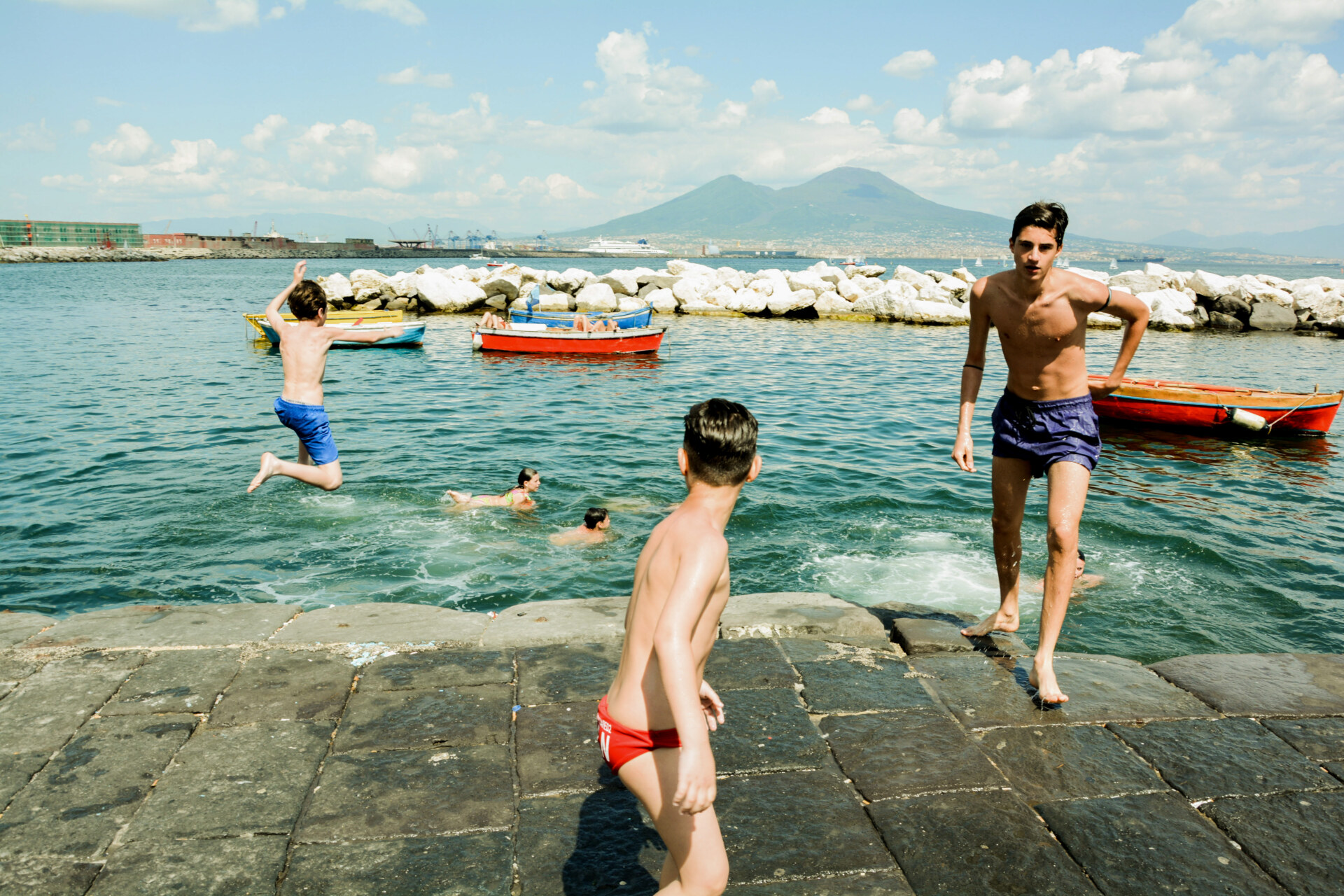 an image by Francesco Freddo of young boys jumping off of the concrete ledge into the clear blue water in the cove off of Naples under Mt Vesuvius