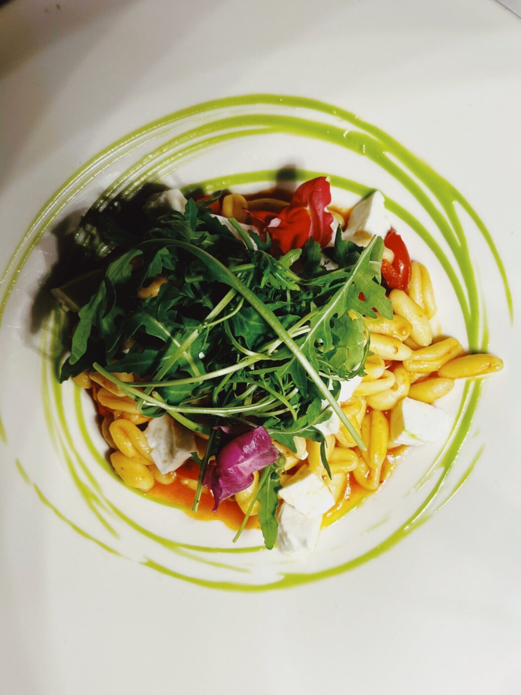 a white bowl with arugula, a grain, feta, and tomatoes and a light green drizzle around it