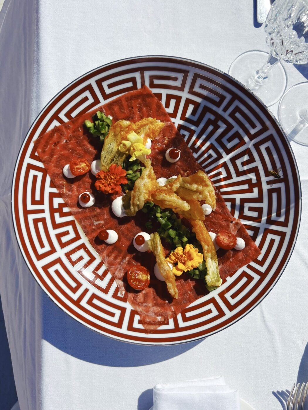 a circular plate with dark red and white geometric lines with a red rectangular food covered in fried vegetables, edible flowers, and dots of a white cream arranged very nicely and specifically