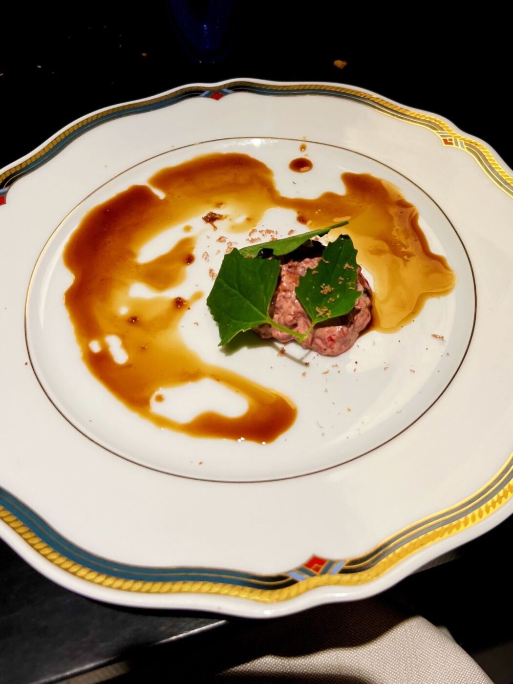 a gold-trimmed white dish with a small scoop of a diced pink meat with three spinach leaves on top, and the rest of the plate is covered with balsamic