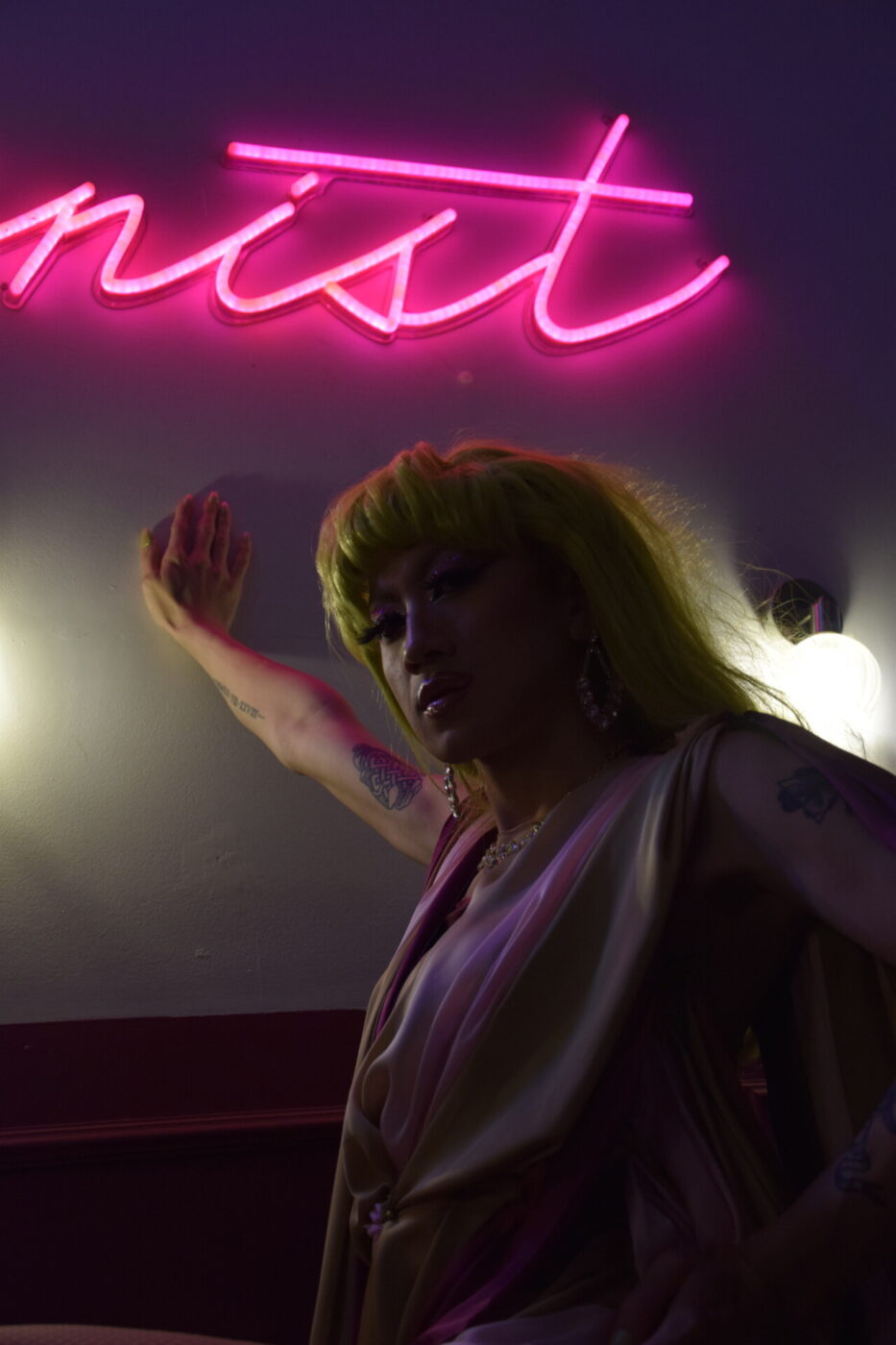 a dark image of a drag queen with bright green hair leaned against the wall under a bright pink neon sign