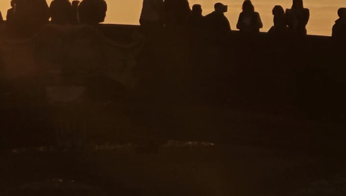 an image of a golden sky above a bridge with dark outlines of couple standing and sitting along it