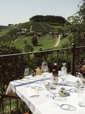 photo by Letizia Cigliutti of a table on a terrace overlooking the rolling hills of monforte d'alba with a bottle of barolo, empty wine glasses, and empty espresso cups