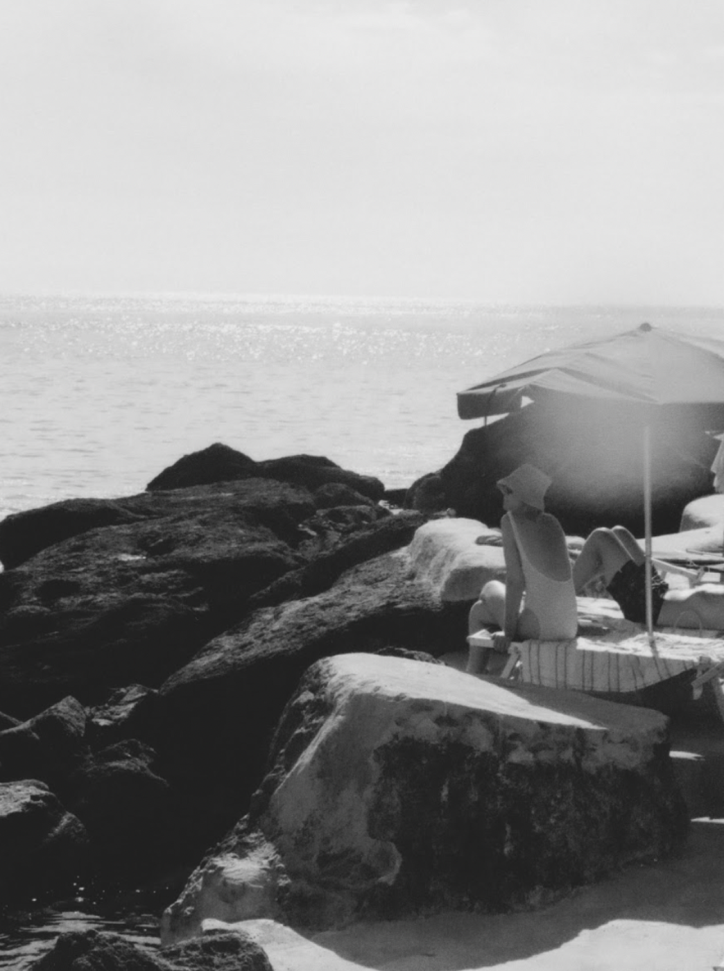a black and white image by Luisa Pagani of two people sunbathing on large rocks near the sea