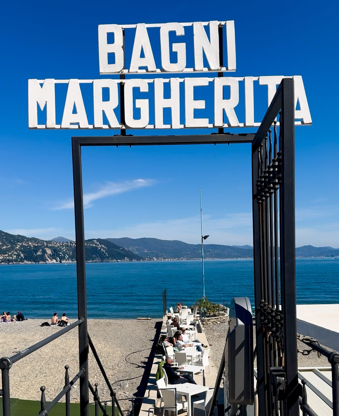 Entrance to Bagni Margherita - Entrance to Bagni Margherita with a view of a pebble beach, blue sea, and clear sky. White chairs and tables are arranged leading to the water. Hills are visible in the background.