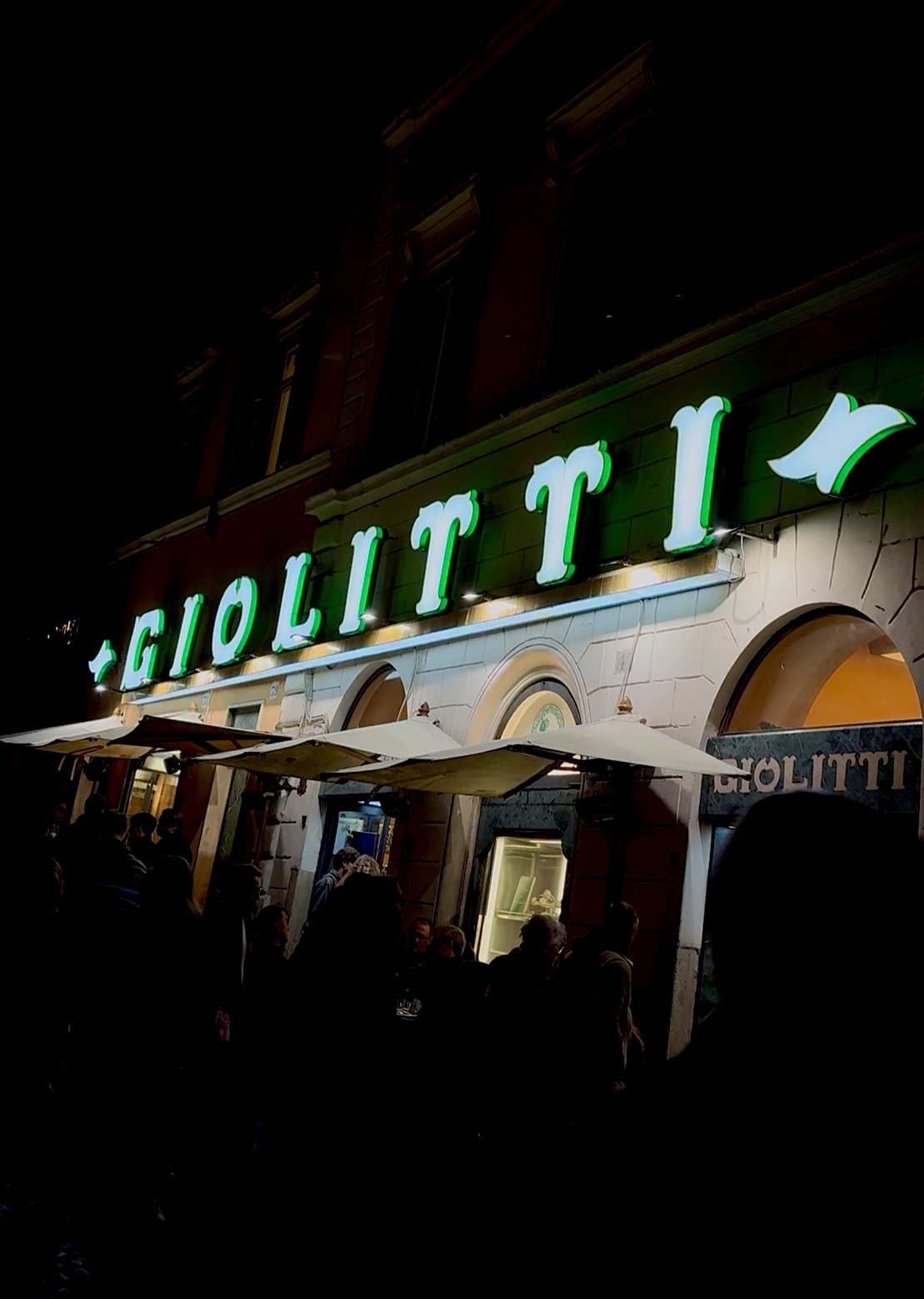 GIOLITTI at Night - A night scene featuring a building with an illuminated neon green sign reading GIOLITTI above an entrance. People are gathered outside under a row of canopies. The background is dark, highlighting the bright sign.