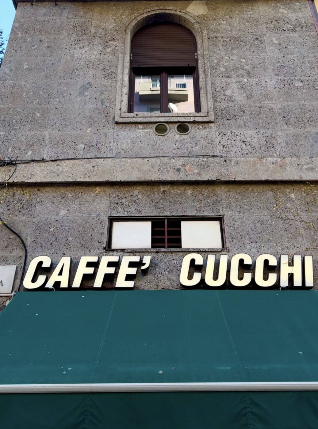 [Image Title] - A stone building facade with a green awning below. The words CAFFÈ CUCCHI are displayed in large, bold, white letters above the awning. A small, arched window is visible at the top of the building.