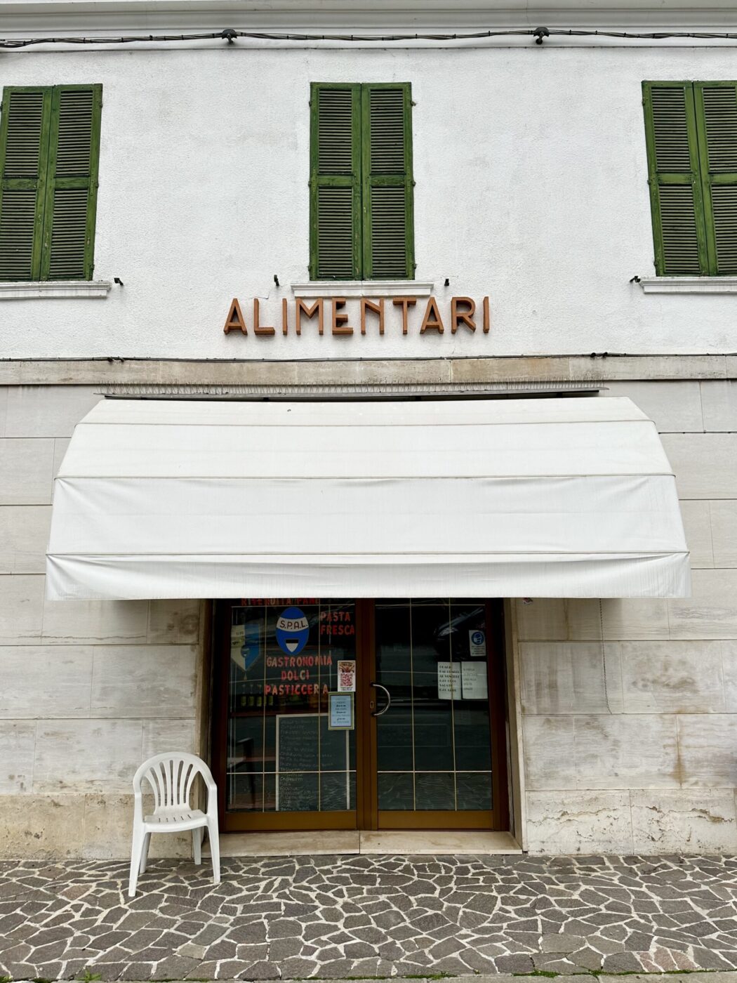 Alimentari Storefront - The image shows the exterior of a small store labeled Alimentari, with green shuttered windows and a white awning above the entrance. A white plastic chair is placed on the stone-paved area outside the store.