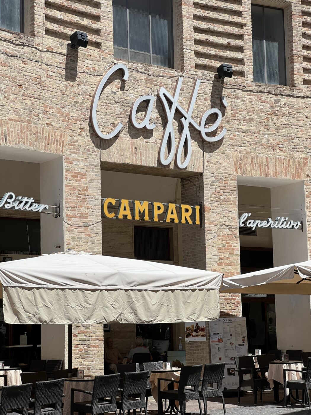 [Image Title] - A brick building with a Caffè sign above a patio area. Signs read Bitter Campari and laperitivo. The patio has tables, chairs, and umbrellas.
