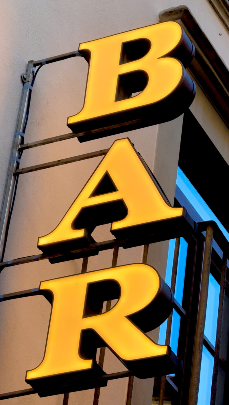 Neon BAR Sign - Bright yellow neon sign displaying the word BAR vertically against a building exterior, with metal brackets securing it to the wall. The letters are outlined for emphasis, and a window is partially visible in the background.
