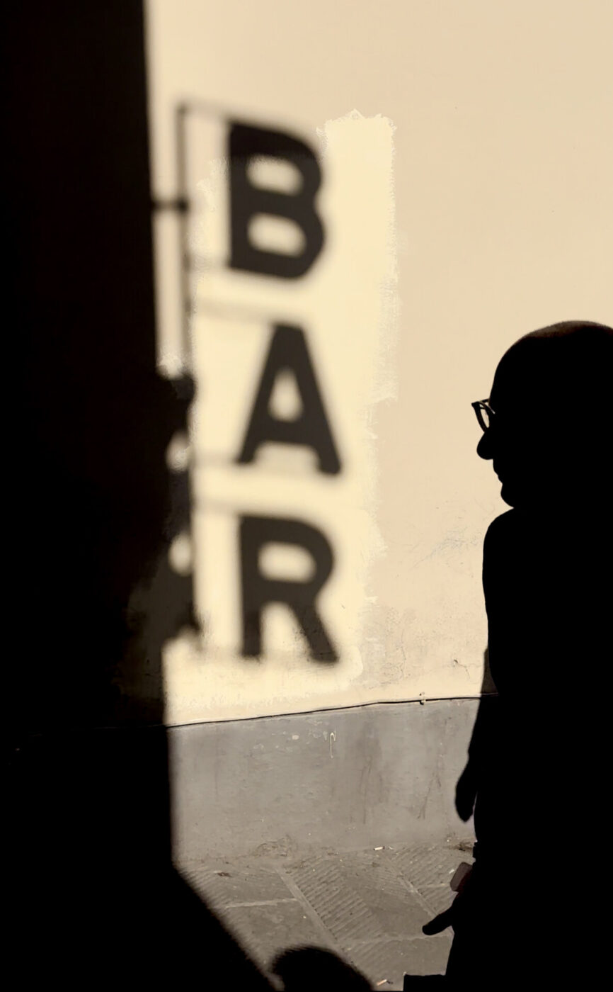 the shadow of the vertical sign for a bar onto a yellow wall and the dark outline of a man walking into the frame