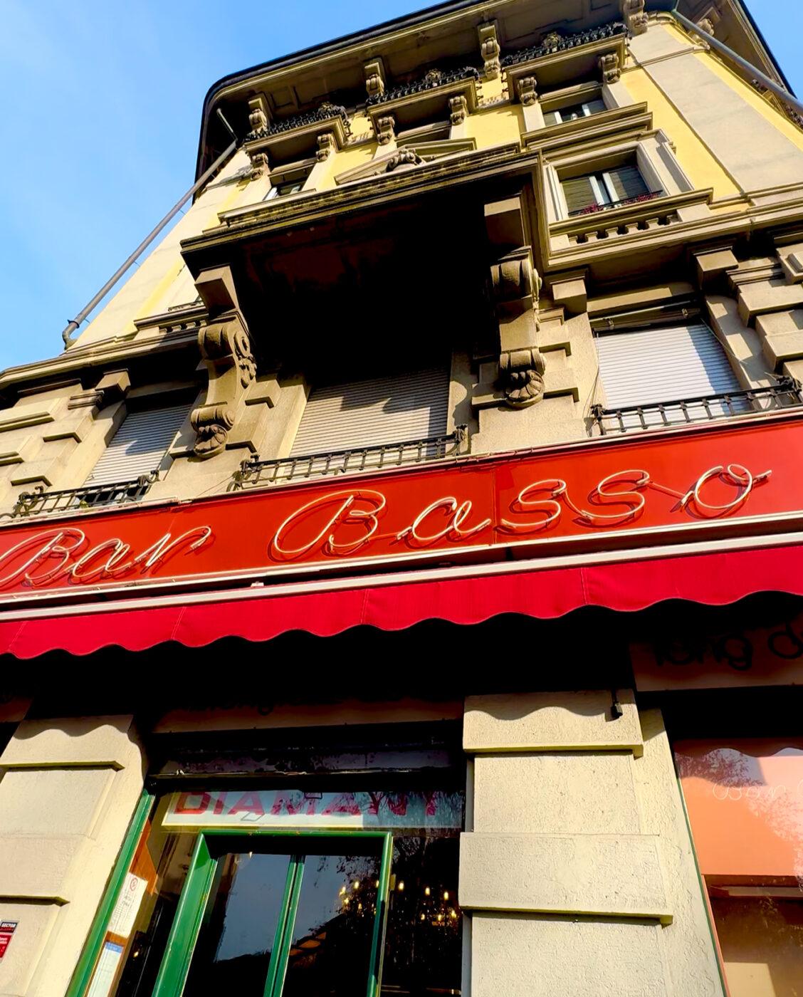 Bar Basso in Milan - A historic building with ornate architecture featuring a red awning. The sign reads Bar Basso in cursive letters. There are large windows on the ground floor with a glimpse of the interior lighting.