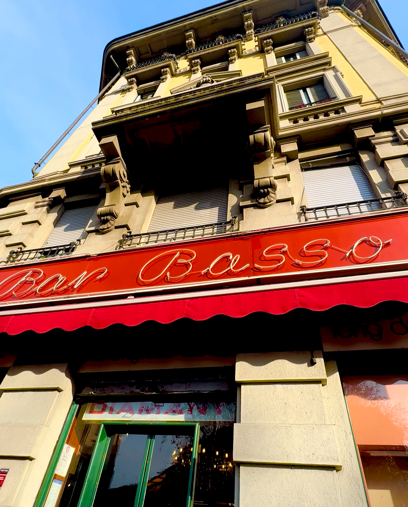 Bar Basso in Milan - A historic building with ornate architecture featuring a red awning. The sign reads Bar Basso in cursive letters. There are large windows on the ground floor with a glimpse of the interior lighting.