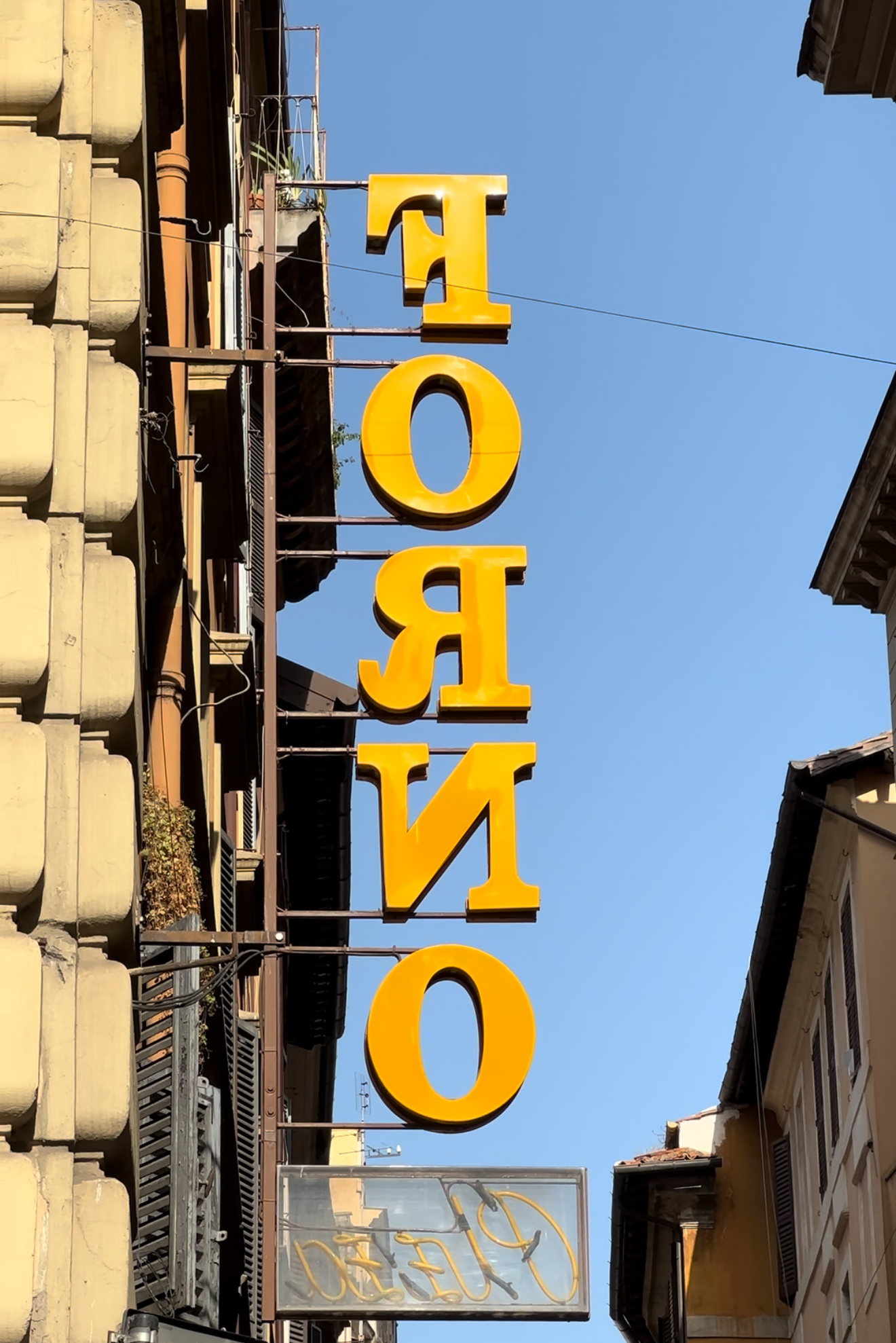 FORNO Sign - A vertical sign displaying the word FORNO in large, yellow letters hangs against a clear blue sky. It is attached to the side of a building with traditional architecture.