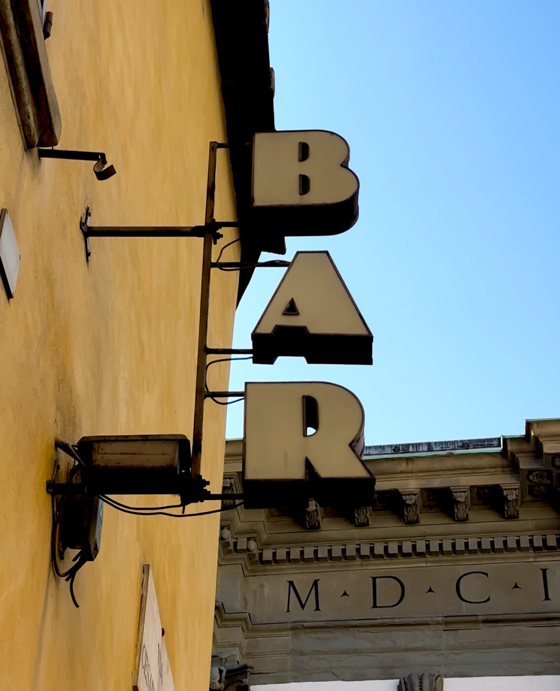 Retro Bar Sign - A retro-style sign with the word BAR in bold letters hangs from the side of a yellow building. An architectural structure with Roman numerals MDCL is partially visible in the background. The sky is clear and blue.