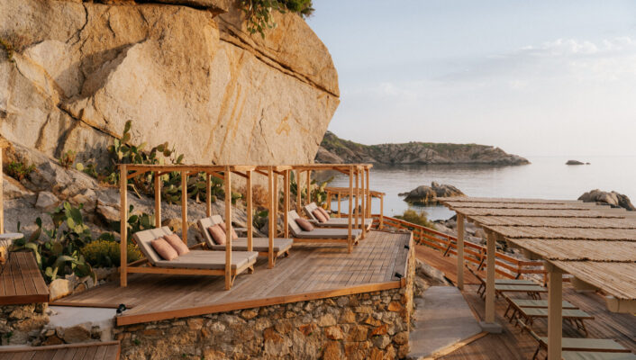 Private wooden cabanas with lounge chairs face a rocky coast by a large formation, blue water, and clear sky.
