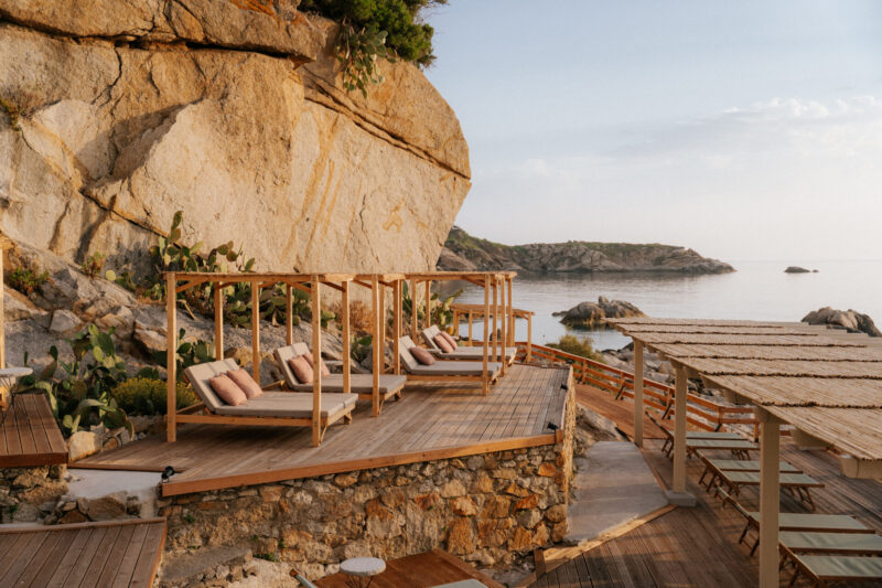 Private wooden cabanas with lounge chairs face a rocky coast by a large formation, blue water, and clear sky.