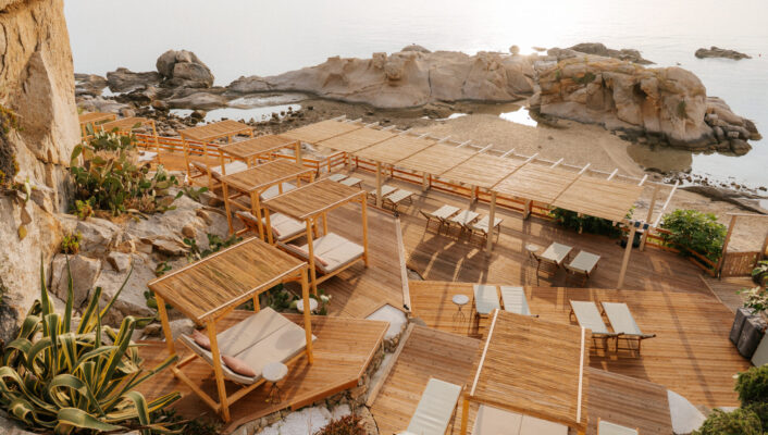 A tranquil beach lounge with wooden cabanas, sunbeds, and pergolas by calm water and rocks at sunrise or sunset.