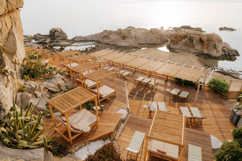 A tranquil beach lounge with wooden cabanas, sunbeds, and pergolas by calm water and rocks at sunrise or sunset.
