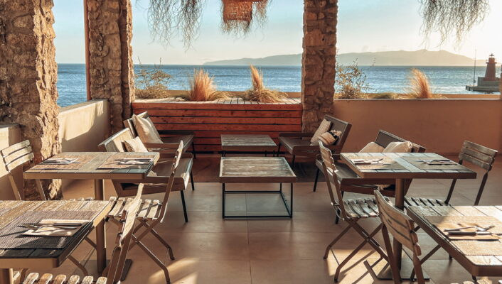 Sunny seaside terrace with wood tables, wicker lights, ocean view through arches, long sun shadows on tile floor.
