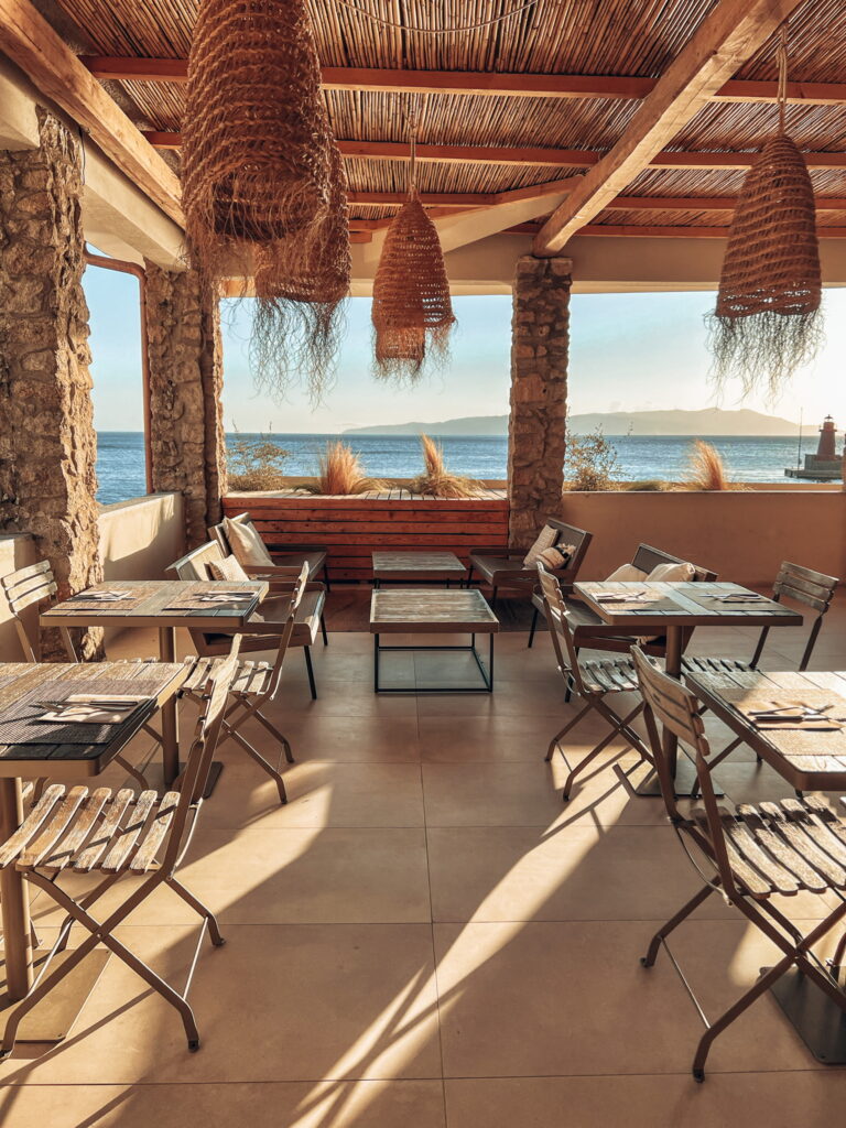 Sunny seaside terrace with wood tables, wicker lights, ocean view through arches, long sun shadows on tile floor.