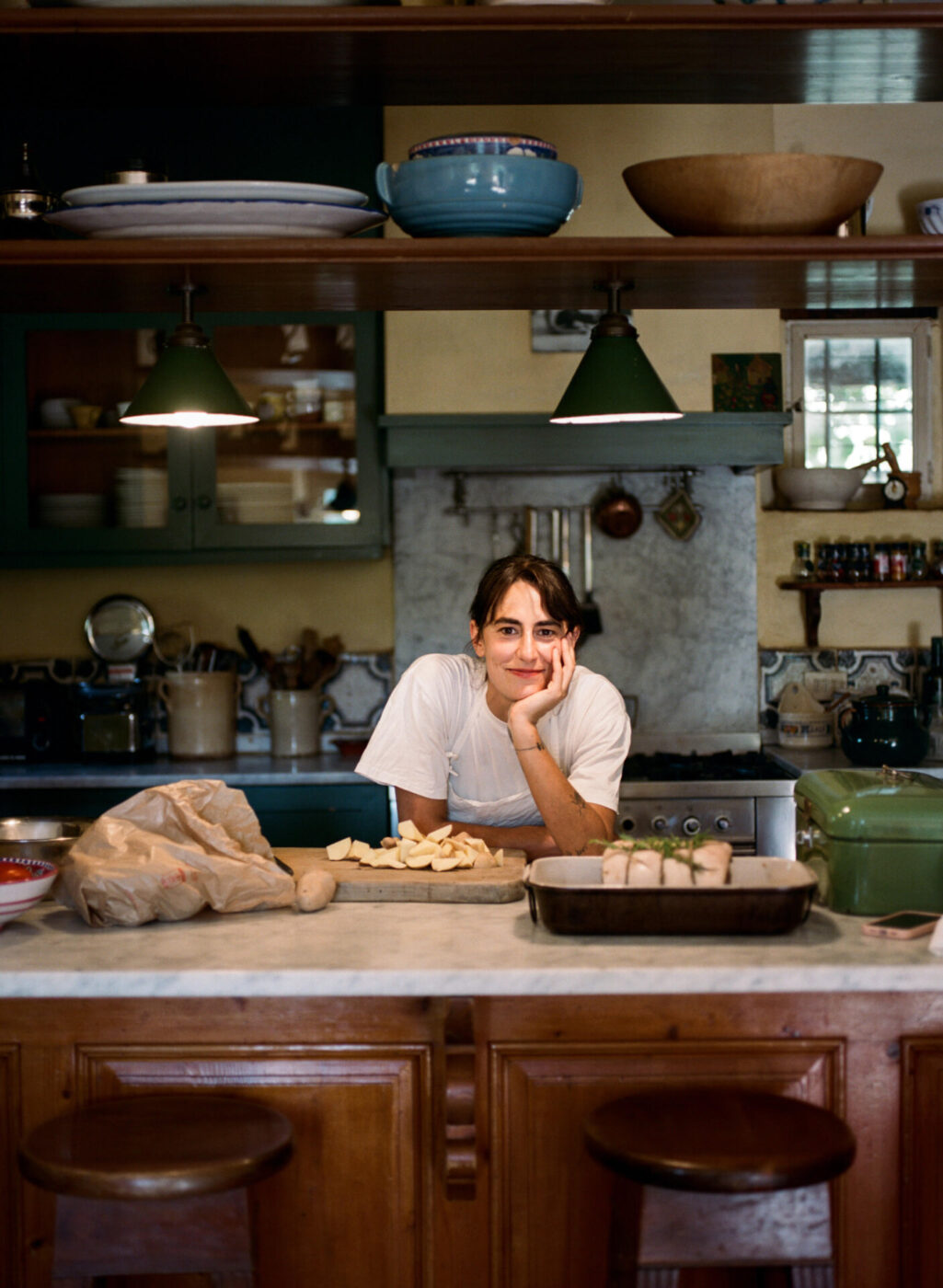 an image of a woman chef resting her heat on her hand behind the counter in the kitchen with porchetta and a cutting board of sliced potatoes