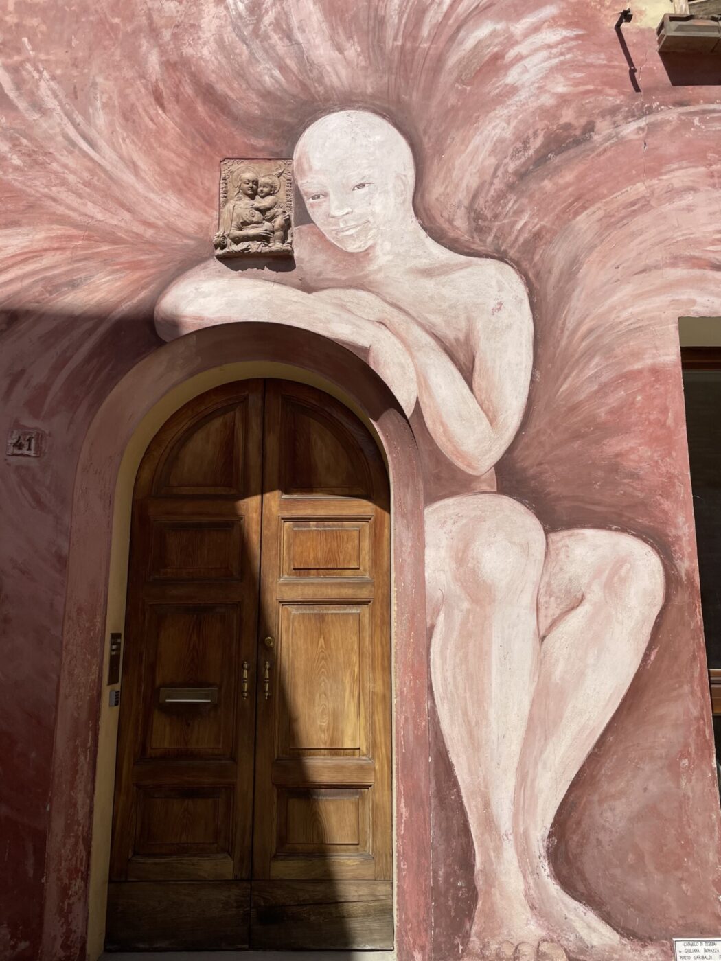 a painted wall in Dozza of a pale pink person leaning on the top of a doorframe with pink all around