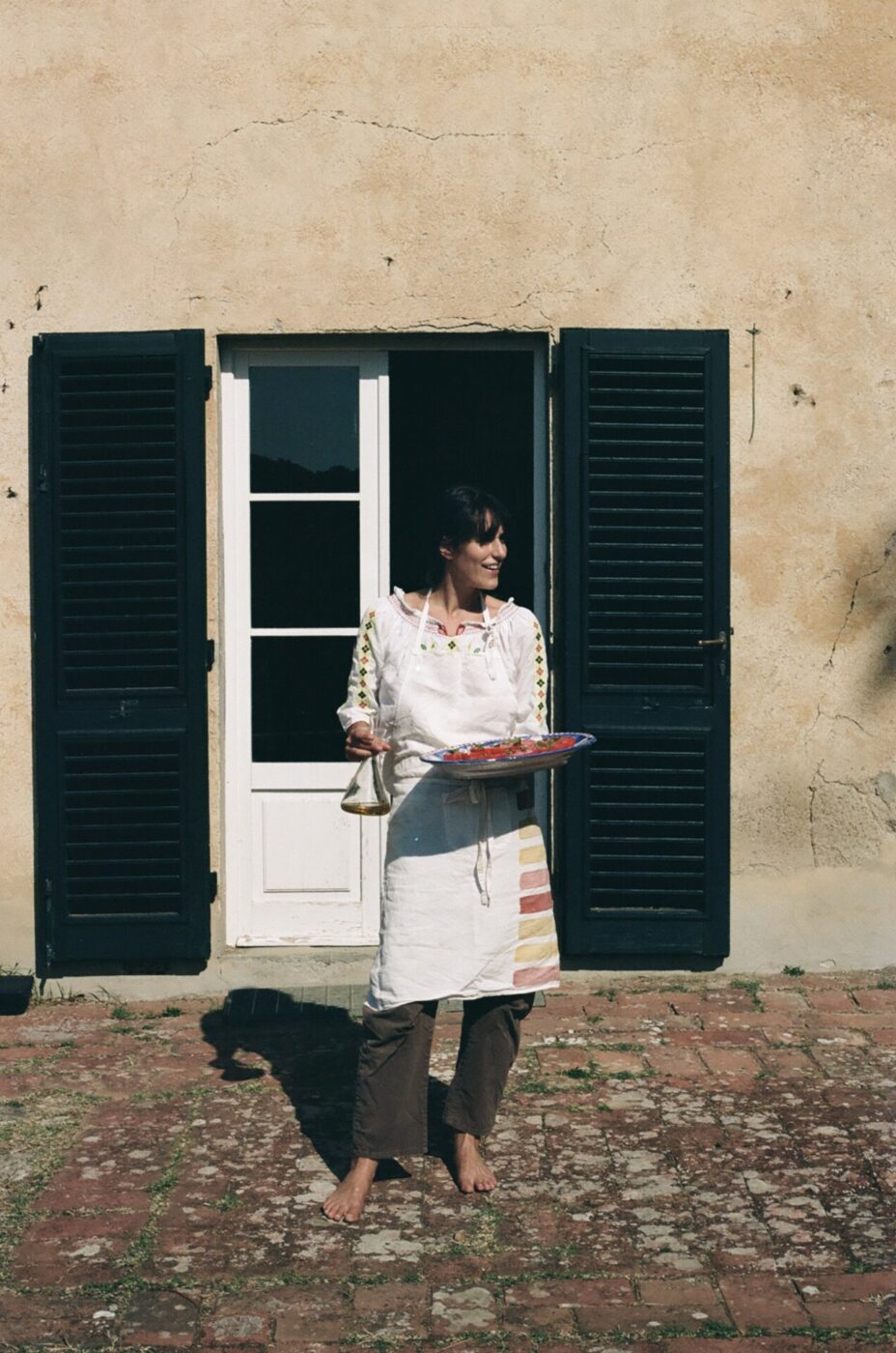 an image of a barefoot woman in a white apron holding a large plate of tomatoes outside a Tuscan farmhouse