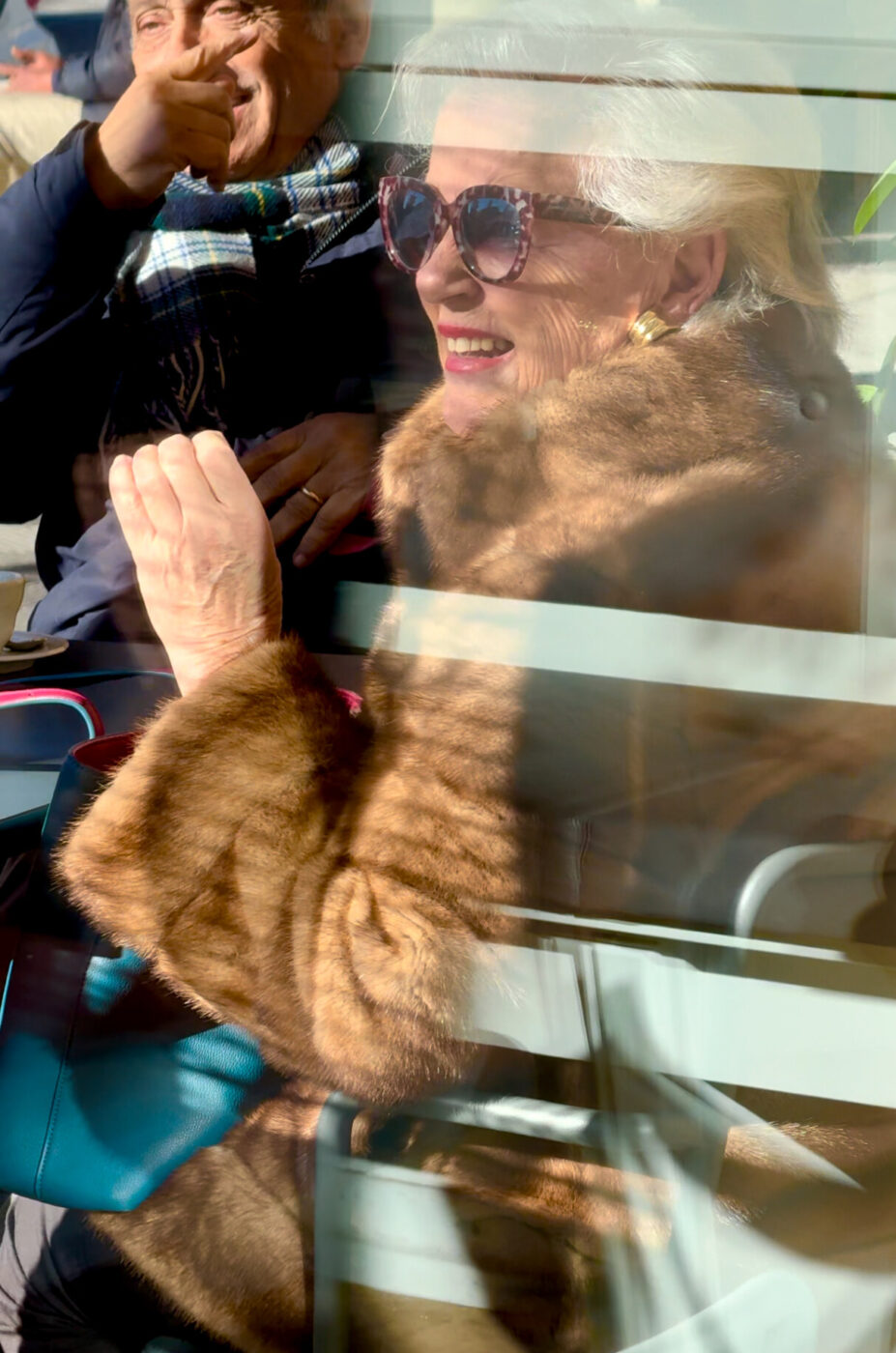 a photo taken through a window of a sciura smiling at a table wearing gold earrings, red lipstick, and leopard print sunglass frames