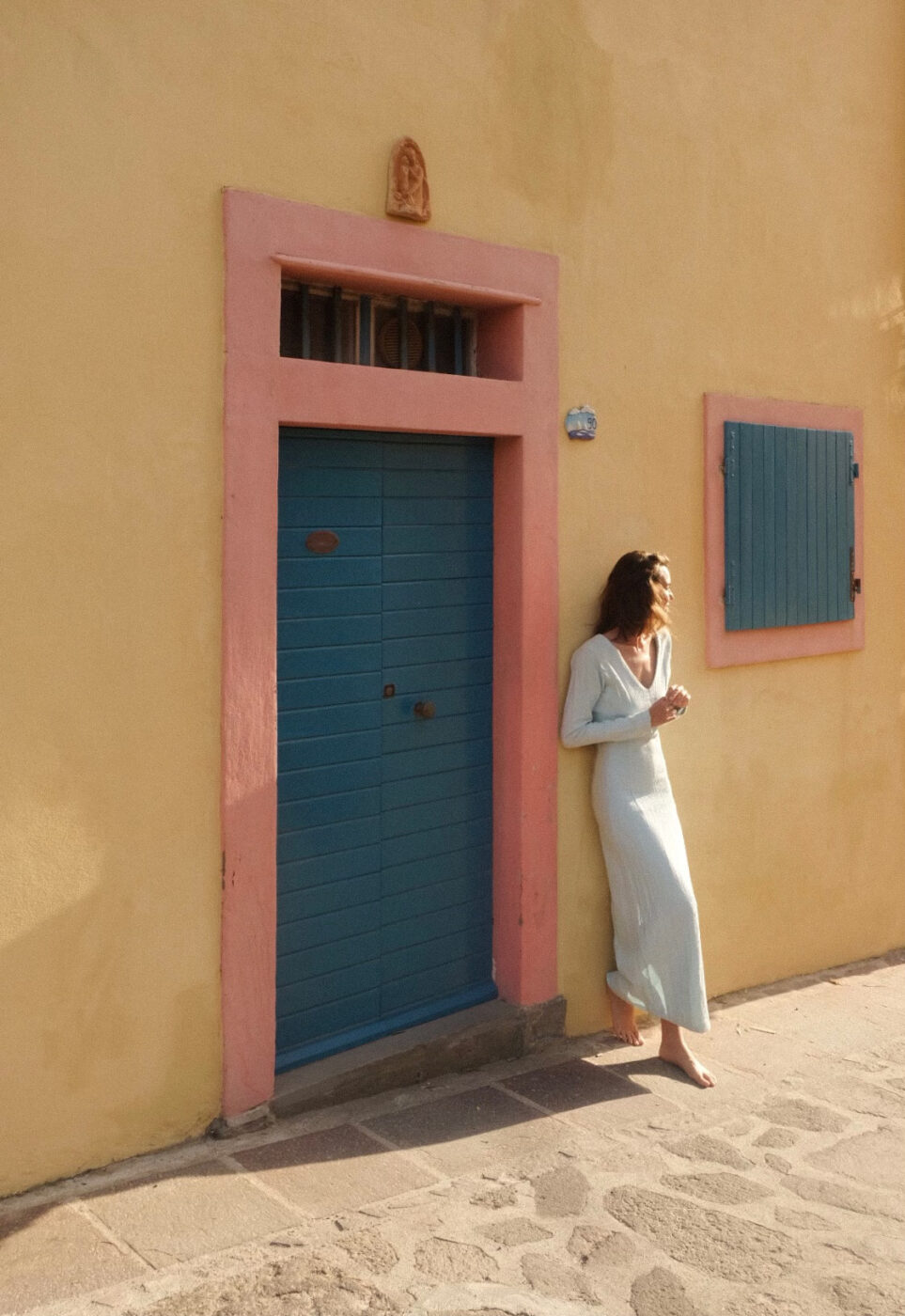 a woman in a long white dress leaning up against a yellow wall near a light blue door with orange trim