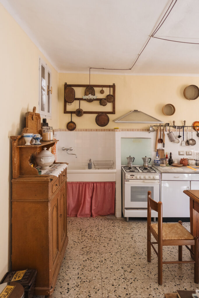 A rustic kitchen with vintage cookware on pale yellow walls, a wooden cabinet, a stove, a sink with a pink skirt, and a small wooden chair on a speckled tile floor.