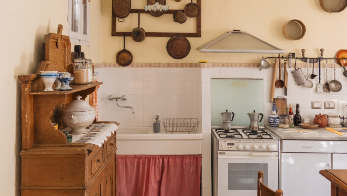 A rustic kitchen with vintage cookware on pale yellow walls, a wooden cabinet, a stove, a sink with a pink skirt, and a small wooden chair on a speckled tile floor.