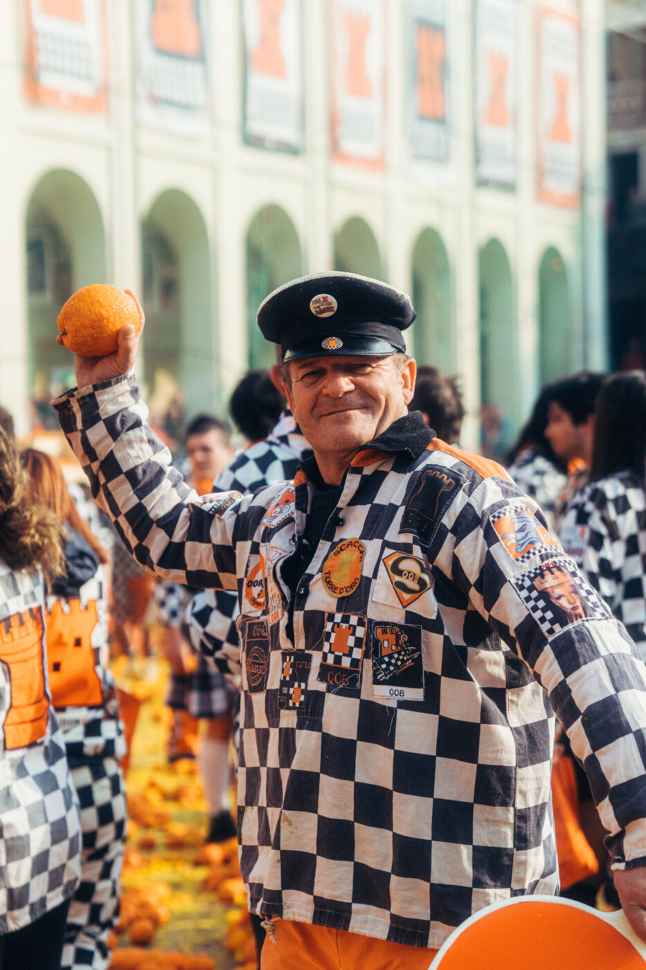 a man in a black and white checkered jacket holding an orange above his head at Ivrea