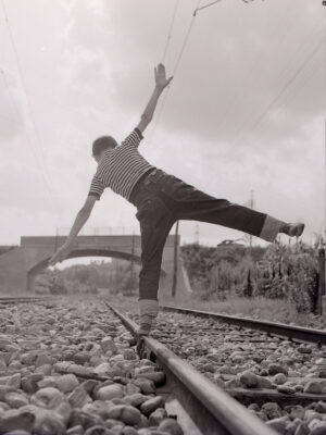 a black and white image of a boy in a striped shirt balancing on one track of a railroad