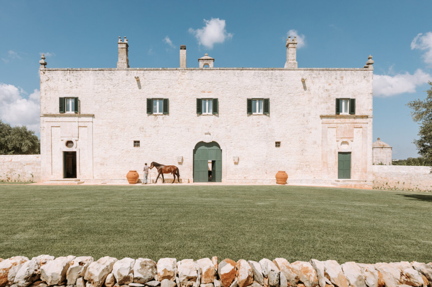 the facade of the stark white, uniform Masseria Lamacoppa with a man walking a horse along the front