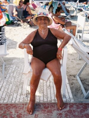 an older tan woman in a brown bathing suit and hat sitting and smiling at a beach club in Puglia