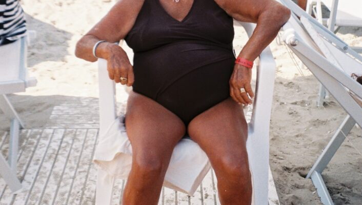an older tan woman in a brown bathing suit and hat sitting and smiling at a beach club in Puglia