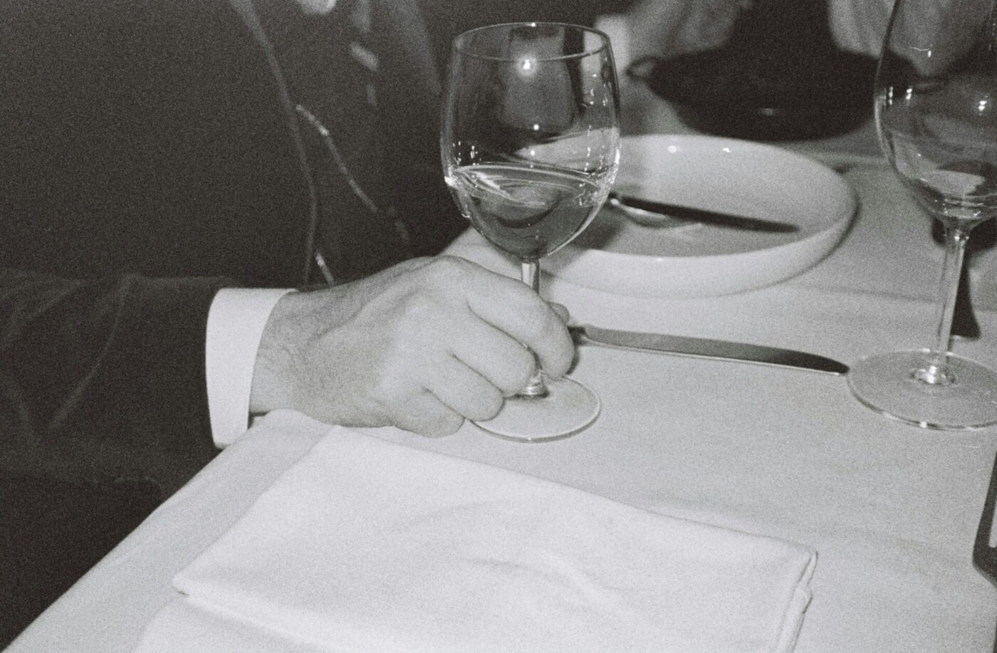 a black and white image of a man holding the stem of a glass of wine at the dinner table