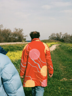 Angelo Milano in a bright orange coat with yellow and pink circles and squiggly lines walking away through farmland in Puglia