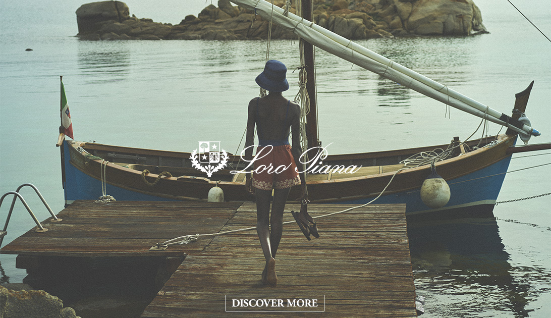 model with blu hat overlooking a vintage wooden fishing boat on dock. Loro Piana brand logo and discover now button ontop