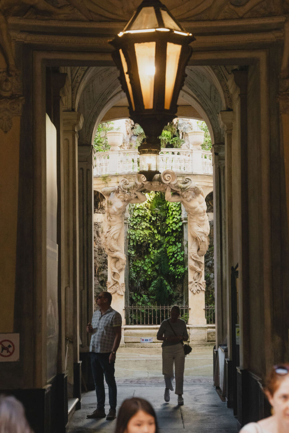 an image through an archway into a courtyard with a statue and greenery in Genova