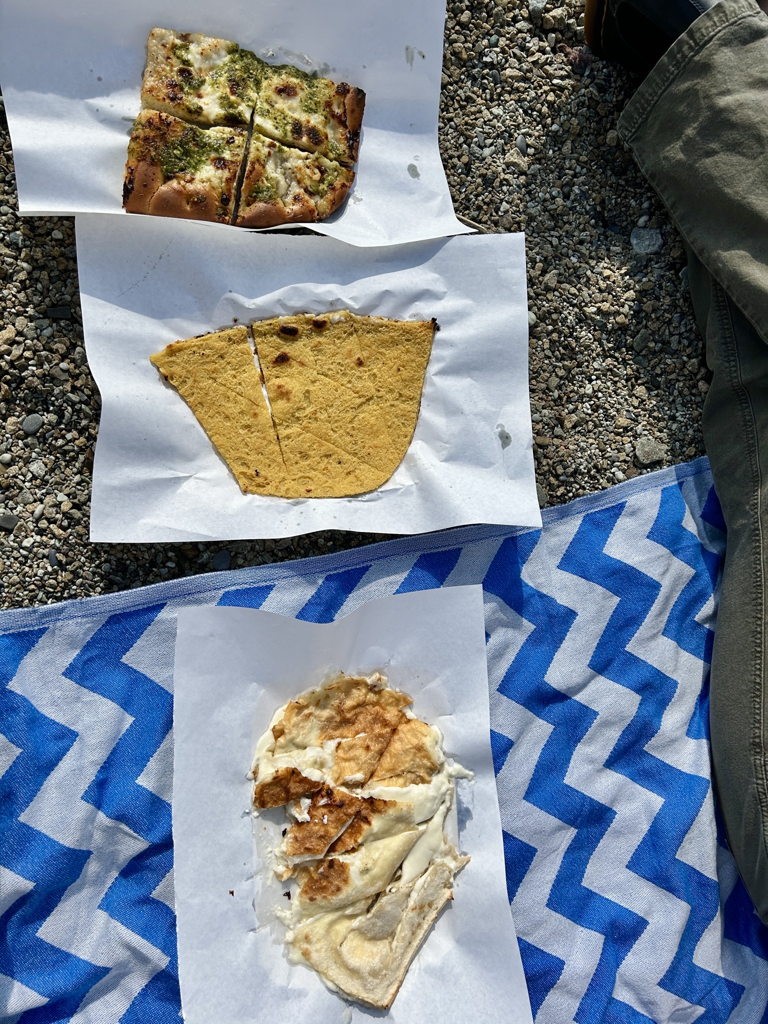 an overhead image of focaccia with pesto, farinata, and focaccia di recco on the beach