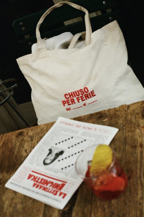 The Estate Italiana Tote Bag - The cream tote bag, emblazoned with red Italian text Chiuso per ferie, sits on a green chair. On the wooden table in front, there is a newspaper and a glass of orange-red cocktail garnished with citrus slices.