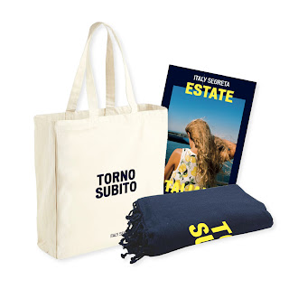 The Estate Italiana Red Bundle - The image shows a beige tote bag with TORNO SUBITO printed on the front, a rolled-up navy blanket, and an ITALY SEGRETA ESTATE magazine that features a woman standing by the sea.