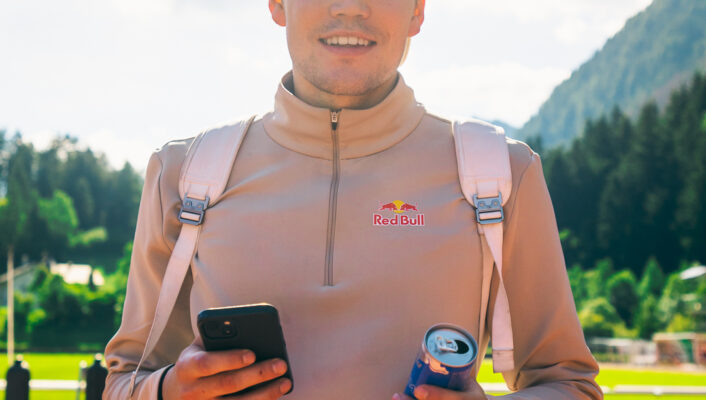an image of a man with a red bull and his phone wearing a backwards hat and a quarter zip ready to go skiing