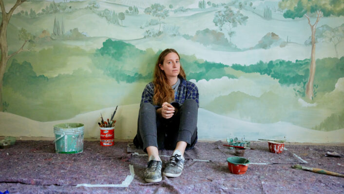 a woman with long dirty blonde hair sitting in front of a mural of rolling green hills