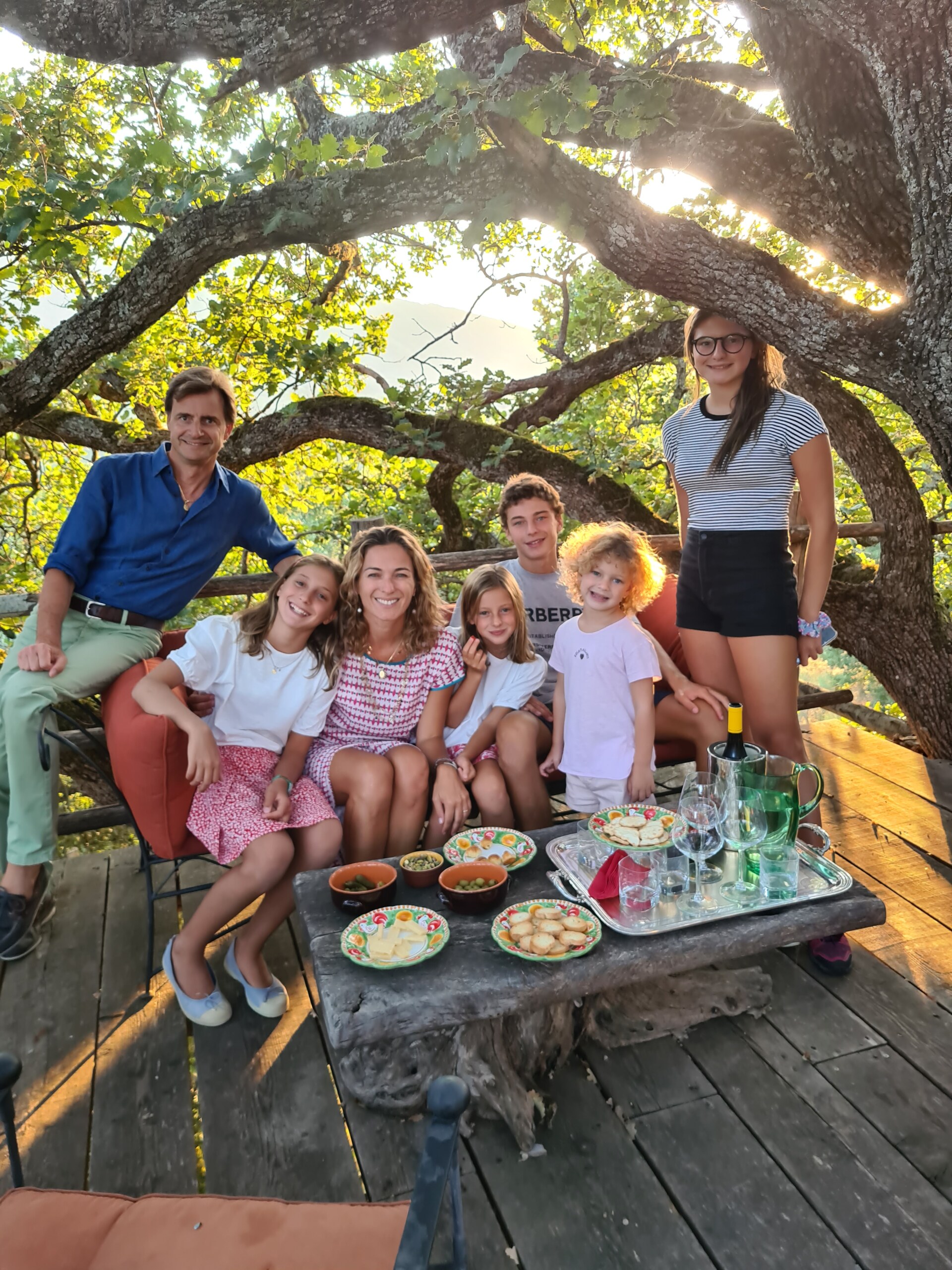a family with five kids sitting outside on a tree-shaded terrace gathered around a small coffee table with snacks and drinks
